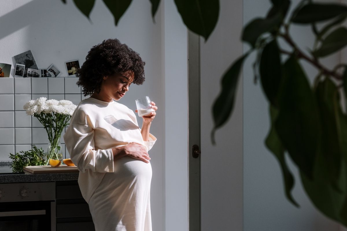 Gestante na cozinha, com a mão na barriga e um copo na outra mão